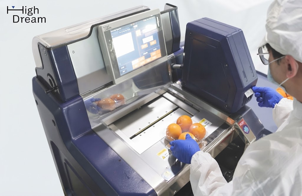 Operator in food-safety clothing using a High Dream weigh-wrap-label machine to wrap and label a tray of fresh citrus in a retail backroom